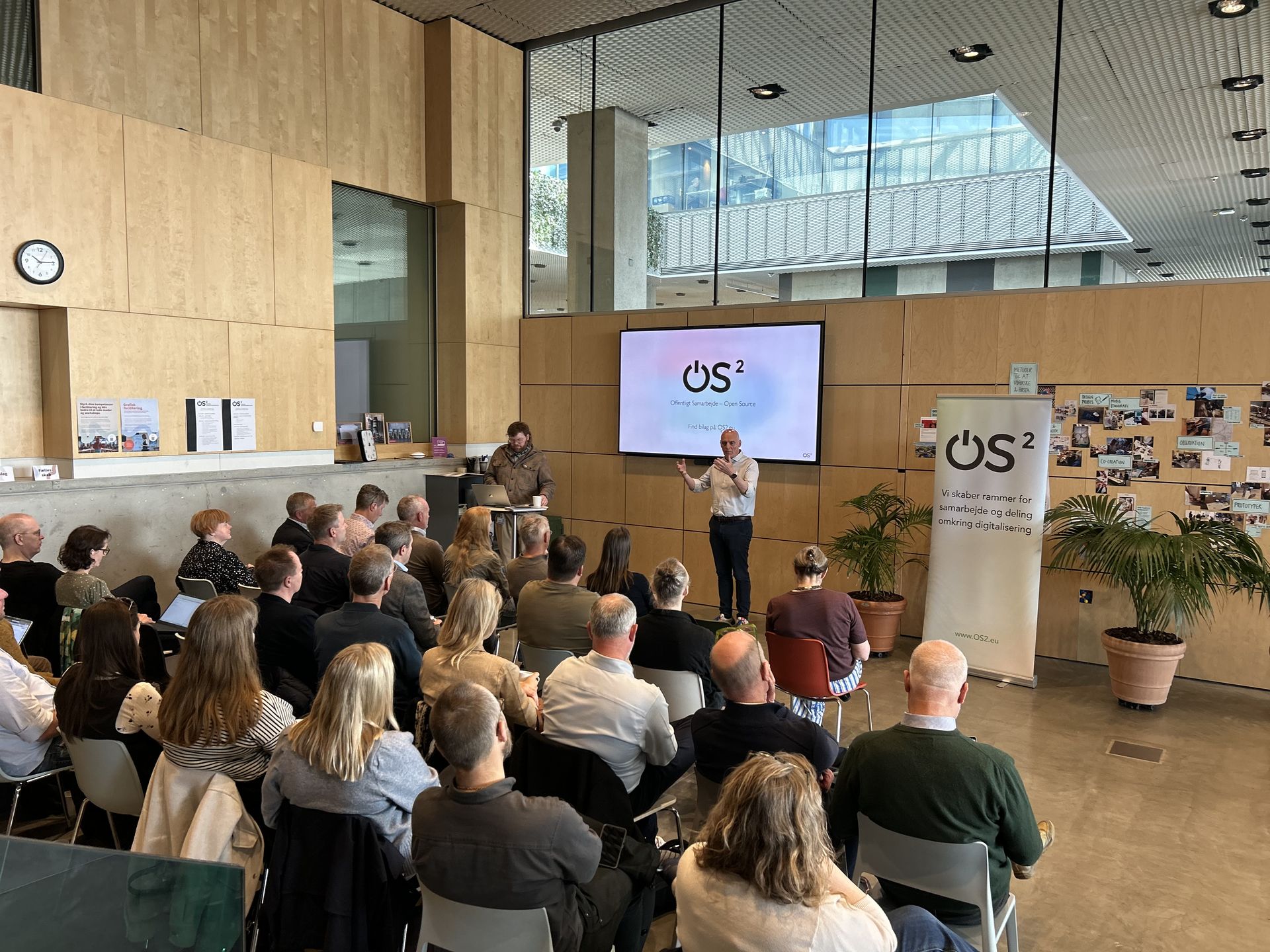 OS2-generalforsamling i Aarhus med oplægsholder, deltagere, storskærm og OS2-banner.